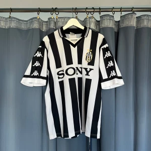 Retro fotbollströja Juventus Kappa - Juventus klassiska fotbollströja från Kappa med svartvita vertikala ränder, broderat klubbmärke och SONY som sponsor. Tröjan har korta ärmar med Kappa-loggor längs sidorna och V-ringad krage. Tillverkad i lätt och ventilerande polyester, perfekt för fotbollsfans. Passar storlek XS-L. Jag är storlek S och köpte den för en oversize look.