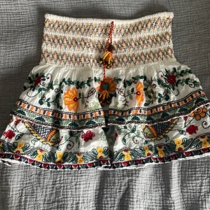 Vit broderad kortkjol med volanger - Superfin vit kortkjol med broderade blommor i gult, rött och grönt. Kjolen har bred smock i midjan, volanger och en dekorativ snörning med tofsar framtill. Perfekt för dig som gillar boho-vibe och färgglada detaljer. Supertrendig från zara! Går ej att köpa längre och supersnygg när man viker ner den!