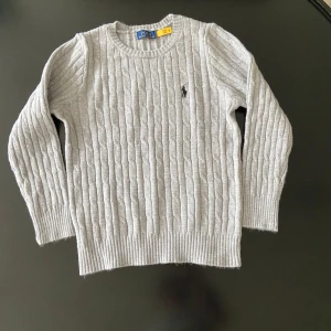 Grå kabelstickad tröja från Polo Ralph Lauren - Kabelstickad grå tröja från Polo Ralph Lauren med rund halsringning och broderad logga på bröstet. Tröjan har långa ärmar och ribbade muddar vid ärmslut och nederkant. Perfekt för lager-på-lager under kyliga dagar.