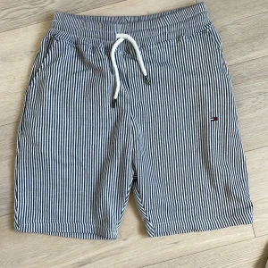 Randiga shorts Tommy Hilfiger M - Snygga randiga shorts från Tommy Hilfiger i vitt och marinblått. De har elastisk midja med snörning, sidofickor och en bakficka. Perfekta för sommaren och tillverkade i mjuk bomull för en chill vibe.