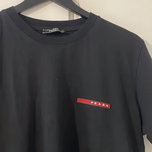 Svart t-shirt från Prada, storlek M - Svart t-shirt från Prada med klassisk passform och korta ärmar. Diskret röd Prada-logga på bröstet ger en clean och exklusiv vibe. Tillverkad i mjuk bomull som känns skön mot huden. Perfekt för dig som gillar stilrena designer och high-end streetwear.