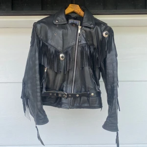 Motorcykel jacka ÄKTA läder - Riktigt cool svart skinnjacka från Grahn's med långa fransar på axlar, rygg och ärmar. Jackan har bälte i midjan, dragkedja framtill och snygga metalldetaljer. Perfekt för dig som vill sticka ut med en edgy och unik stil.