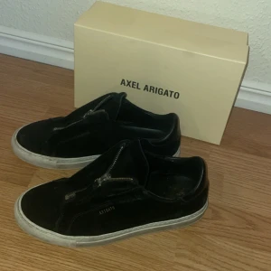 Svarta sneakers från Axel Arigato - Snygga svarta sneakers från Axel Arigato med dragkedja framtill. Skorna har en stilren design med vit sula och är perfekta för en casual look.