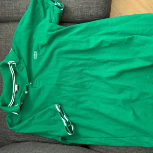 Grön pikétröja från Lacoste - Säljer en klassisk grön pikétröja från Lacoste med den ikoniska krokodilloggan på bröstet. Tröjan har korta ärmar och en krage med vita detaljer. Perfekt för en stilren och sportig look.
