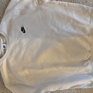 Vit sweatshirt från Nike - Säljer en stilren vit sweatshirt från Nike med deras klassiska logga i svart på bröstet. Tröjan har långa ärmar och är tillverkad i ett mjukt material som passar perfekt för en avslappnad stil. Perfekt för både vardag och träning. Storlek M. Mycket bra skick