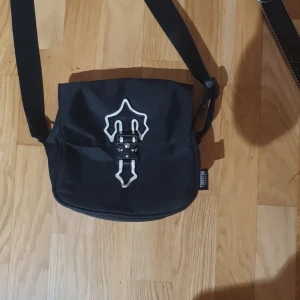 Svart axelväska från Trapstar - Snygg svart axelväska från Trapstar med ett unikt vitt mönster på framsidan. Väskan har en justerbar axelrem och en praktisk ficka med dragkedja. Perfekt för att bära med sig det viktigaste med stil.