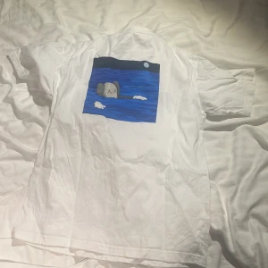 Sweat KAWS - Säljer en vit t-shirt med ett unikt konstmotiv på baksidan i blått och grått. Framsidan har en liten detalj i samma stil. Perfekt för dig som gillar konstnärliga tryck!
