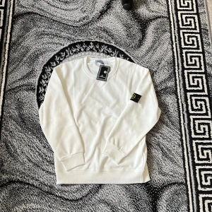 Vit sweatshirt från Stone Island - Säljer en stilren vit sweatshirt från Stone Island med deras ikoniska märke på ärmen. Tröjan har en klassisk rund hals och ribbade muddar vid ärmslut och nederkant. Perfekt för en avslappnad look.