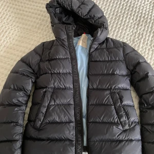 Mörkblå dunjacka från Moncler storlek 152  - Säljer en snygg mörkblå övergångs dunjacka modell anni från Moncler med huva och dragkedja. Jackan har en stilren design med Moncler-logga på dragkedjan och en ljusblå insida. Perfekt för vapen och kalla sommardagar. Jackan är i mycket bra skick lätt figursydd modell storlek 152 ca 12 år.