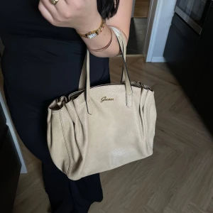 Beige handväska från Guess - Snygg beige vintage handväska från Guess med dragkedja och logotypdetaljer. Perfekt för att bära dina dagliga nödvändigheter med stil. Väskan har ett rymligt inre med mönstrat foder och flera fack för enkel organisering. Tyvärr syns användning på den där man håller i, men annars fint skick!