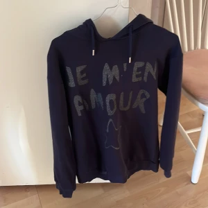Mörkblå hoodie med text - Säljer en mörkblå hoodie med texten 'JE M'EN AMOUR' i glittrigt tryck på framsidan. Tröjan har en klassisk huva med dragsko och långa ärmar. Perfekt för en avslappnad stil. Det finns en liten fläck som jag inte har testat att ta bort.