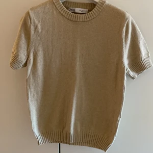 Beige kortärmad tröja från Zara - Säljer en stilren beige tröja från Zara med korta ärmar och ribbade detaljer vid hals, ärmslut och nederkant.  Storlek s men passar mer xs