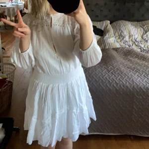 Vit klänning med spetsdetaljer - Charmig vit klänning med spetsdetaljer och långa ärmar. Den har en smockad midja och en luftig kjol. Använd en gång