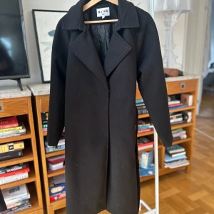 Svart kappa med knytskärp från NA-KD - Säljer en stilren svart kappa från NA-KD i storlek 34. Kappan har en klassisk design med långa ärmar och ett bälte i midjan för en elegant siluett. Perfekt för höst och vår. Ett måste i garderoben för en chic look!