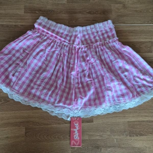 Rutig rosa kjol från Romwe - Söt rosa och vit rutig kjol i tunnt material från Romwe med volangdetaljer och spetskant. Tillverkad i 100% bomull för en bekväm känsla. Perfekt för en somrig look! Storleken är uppskattad till M. Märkning saknas. Aldrig använd.