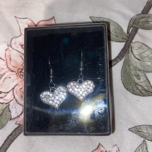 Glittriga hjärtformade örhängen - Snygga örhängen i form av hjärtan med glittriga stenar. Perfekta för att ge en extra touch av glamour till din outfit. De är i en elegant silverfärg och kommer i en fin förpackning., oanvända 