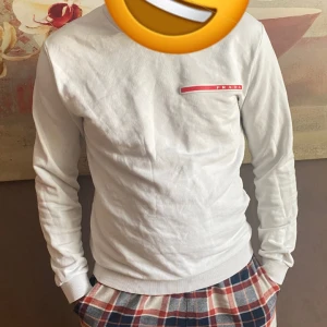 Vit tröja från Prada - Säljer en stilren vit tröja från Prada med en röd logga på bröstet. Tröjan har långa ärmar och ribbade muddar vid ärmslut och nederkant. Perfekt för en avslappnad och trendig look.