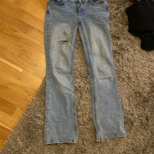 Blå jeans med slitningar - Säljer ett par klassiska blå jeans med slitningar på framsidan. De har en dragkedja och knapp framtill samt fem fickor. Perfekta för en avslappnad stil.