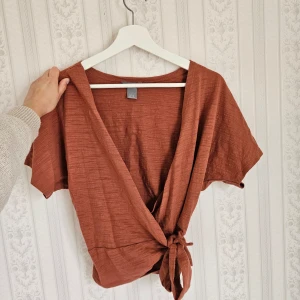 Omlottopp - Säljer en snygg rostfärgad omlottopp med korta ärmar och knytning framtill. Perfekt för fest & vardag 🩷 Endast använd en gång - som ny