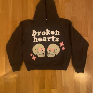 Brun hoodie med tryck (Broken-Planet) - Säljer en brun hoodie med texten 'broken hearts' och ett tryck av två dödskallar och rosa stjärnor på framsidan. Perfekt för en avslappnad stil.