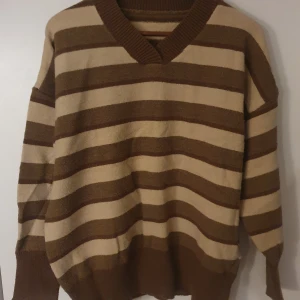 Brun och beige randig tröja - Säljer en mysig brun och beige randig tröja med v-ringning. Perfekt för kyliga dagar med sin avslappnade passform och långa ärmar. 