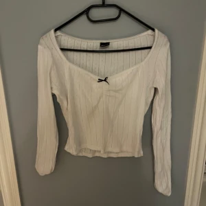 Vit topp från Gina Tricot - Säljer en söt vit ribbad topp från Gina Tricot med en liten svart rosett framtill. Toppen har långa ärmar och en v-ringad design. Perfekt för en stilren look. Skriv gärna om ni har några frågor! 