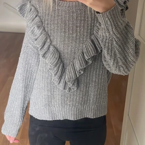 Grå stickad tröja med volangdetaljer - Säljer en snygg grå stickad tröja med volangdetaljer framtill. Tröjan har långa ärmar och en bekväm passform. Perfekt för kyligare dagar!
