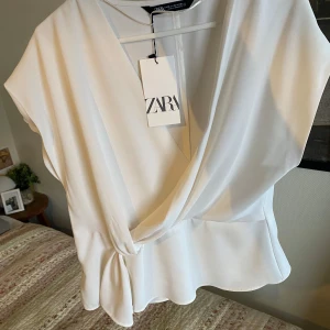 Vit omlottopp från Zara - Säljer en stilren vit omlottopp från Zara. Toppen har en elegant v-ringning och knytning i sidan som ger en fin detalj. Perfekt för en chic look! DET ÄR INGEN FLÄCK PÅ TRÖJAN DE ÄR MIN KAMERA LENS SOM HAR EN FLÄCK