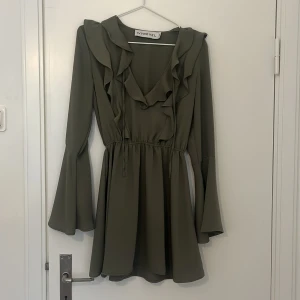 Olivgrön klänning från Ivyrevel - Snygg olivgrön klänning från Ivyrevel med volangdetaljer och långa ärmar. Klänningen har en v-ringad design och knytband i midjan för en smickrande passform. Perfekt för en stilren look.