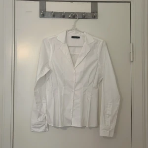 Vit skjorta från Stockh lm  - Elegant vit skjorta från Stockh lm med klassisk krage och knappar framtill. Skjortan har långa ärmar och en figurnära passform som ger en stilren look. Perfekt för både formella och avslappnade tillfällen.