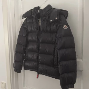 Svart dunjacka från Moncler - Snygg svart dunjacka från Moncler med huva och dragkedja. Jackan har ett quiltat mönster och Moncler-logga på ärmen. Perfekt för kyliga dagar.🤩