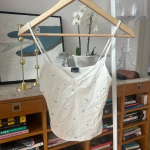 Vit linnetopp från Gina Tricot - Söt vit linnetopp från Gina Tricot med små blå blommor. Tillverkad i en mjuk blandning av bomull och modal. Perfekt för varma dagar med sina smala axelband och enkla design.