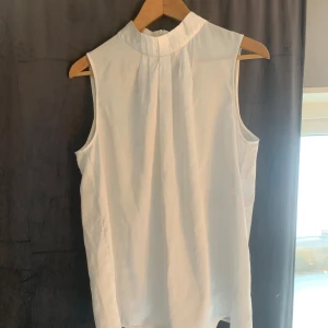 Vit ärmlös blus från Stockh LM - Säljer en stilren vit ärmlös blus från Stockh LM. Blusen har en hög krage och ett elegant fall. Perfekt för en chic och minimalistisk look. Passar bra till både jeans och kjol.