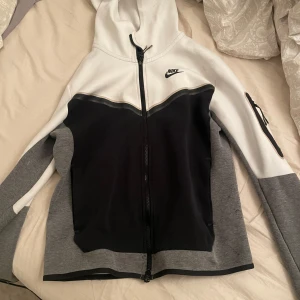 Vit och grå hoodie från Nike tech  - Säljer en snygg vit och grå hoodie från Nike med dragkedja och huva. Jackan har en cool design med svarta detaljer och en praktisk ficka med dragkedja på ärmen. Perfekt för en sportig look!