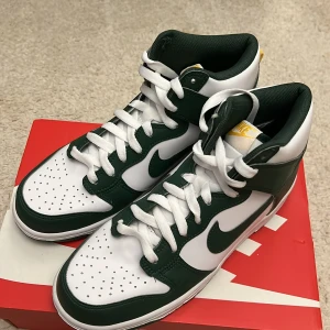 Nike Dunk High GS - Snygga Nike Dunk High sneakers i en fräsch grön och vit färgkombination. Skorna har klassisk snörning och en hög siluett som ger extra stöd. Perfekta för dig som vill ha en stilren och sportig look.