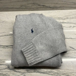 Grå stickad tröja från Polo Ralph Lauren - Säljer en stilren grå stickad tröja från Polo Ralph Lauren med dragkedja och ribbad krage. Tröjan har ett broderat logotyp på bröstet. Perfekt för kyliga sommardagar!