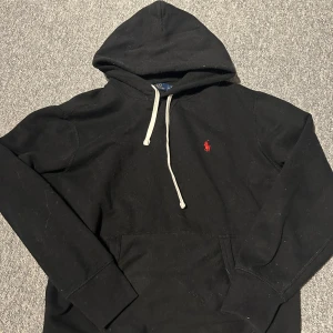 Svart hoodie från Ralph Lauren - Säljer en klassisk svart hoodie från Ralph Lauren med en röd logga på bröstet. Den har en justerbar huva med vita snören och en stor ficka framtill. Perfekt för en avslappnad stil.
