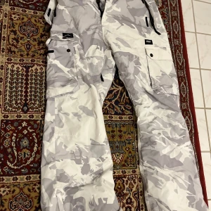 Dope skidkläder byxor och jacka - Snygga vita camo täckbyxor från Dope med flera praktiska fickor och justerbar midja. Perfekta för vinterns äventyr med hög vattentäthet och andningsförmåga. Byxorna har en modern look med camouflagemönster i grått och vitt.Kostar 3800 nytt. Använt 3 tillfällen 