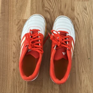 Adidas fotbollsskor i rött och vitt - Snygga Adidas fotbollsskor i rött och vitt med klassiska tre ränder på sidan. Skorna har en färgglad sula i gult och rött och är utrustade med snörning för bästa passform.