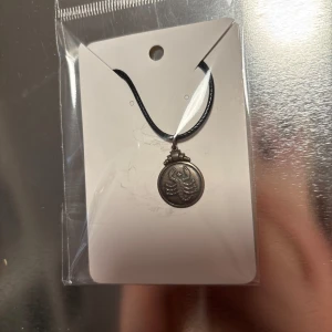 Silverfärgat halsband med skorpionhänge - Snyggt halsband med ett silverfärgat skorpionhänge. Hänget är runt och hänger i en svart rem. Perfekt för att ge en edgy touch till din stil. 