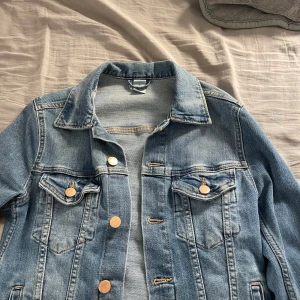 Blå jeansjacka  - Säljer en klassisk blå jeansjacka. Jackan har en normal passform med långa ärmar och knappar framtill. Perfekt för vår och sommar!