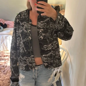 Svart bomberjacka med djurmönster - Säljer en cool svart bomberjacka med ett unikt djurmönster i vitt. Jackan har dragkedja framtill och långa ärmar. Perfekt för att ge din outfit en edgy touch! Har annons med både storlek 34 och 36. 