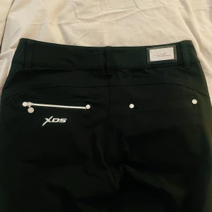 Svarta, lågmidjade byxor från XDS - Snygga svarta byxor från XDS med silverfärgade detaljer och dragkedja på bakfickan. Byxorna har en stilren design med två knappar framtill och en metallplatta med logotyp på baksidan. Perfekta för en stilren look.