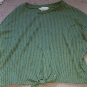 Grön ribbad topp från H&M - Snygg grön ribbad topp från H&M med knytning framtill. Perfekt för en avslappnad stil. Den har en rund halsringning med långa ärmar.