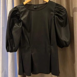 Svart topp med puffärmar med en tajt waist line - Snygg svart topp från Gina Tricot med puffiga trekvartsärmar. Tillverkad i ett glansigt lack material som ger en lyxig känsla. Perfekt för att lyfta vilken outfit som helst. 🖤