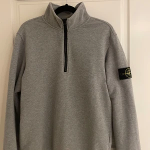 Grå hafe zip från Stone Island - Säljer en stilren grå tröja från Stone Island med halv dragkedja och deras ikoniska märke på ärmen. Perfekt för en avslappnad och trendig look. Tröjan har långa ärmar och en hög krage.