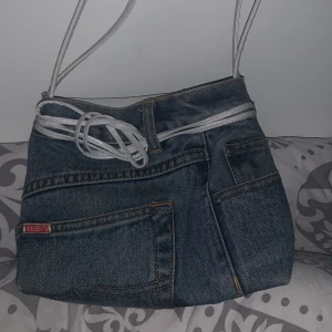 Jeansväska med vita axelband - Säljer en unik jeansväska med vita axelband och en cool design som liknar ett par jeans. Väskan har en ficka framtill och en dekorativ vit snörning. Perfekt för en avslappnad stil!