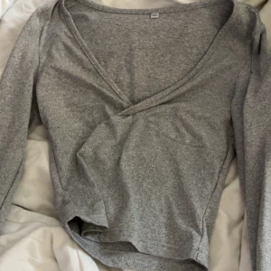 Grå omlottopp med v-ringning XXS - Grå långärmad topp i omlottmodell med djup v-ringning. Tunn och mjuk, perfekt för lager-på-lager. Enkel och stilren design som passar till jeans eller kjol. Modellen är figurnära och har en klassisk look.