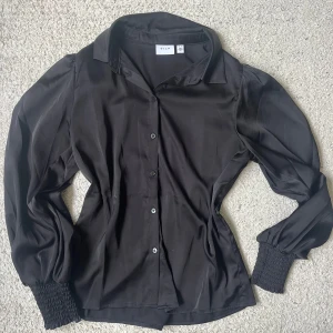 Svart blus från Vila med puffärm - Snygg svart blus från Vila med långa puffärmar och breda smockade muddar vid handlederna. Blusen har klassisk krage och knäppning framtill. Materialet är glansigt och känns lyxigt, perfekt för att lyfta din outfit.
