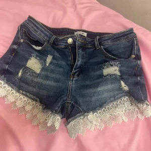 Jeansshorts med spetskant - Snygga blå jeansshorts med slitna detaljer och vit spetskant längst ner. Klassisk femficksmodell med dragkedja och knapp framtill. Perfekta för sommaren och ger en söt men edgy vibe med den rivna looken och spetsen.